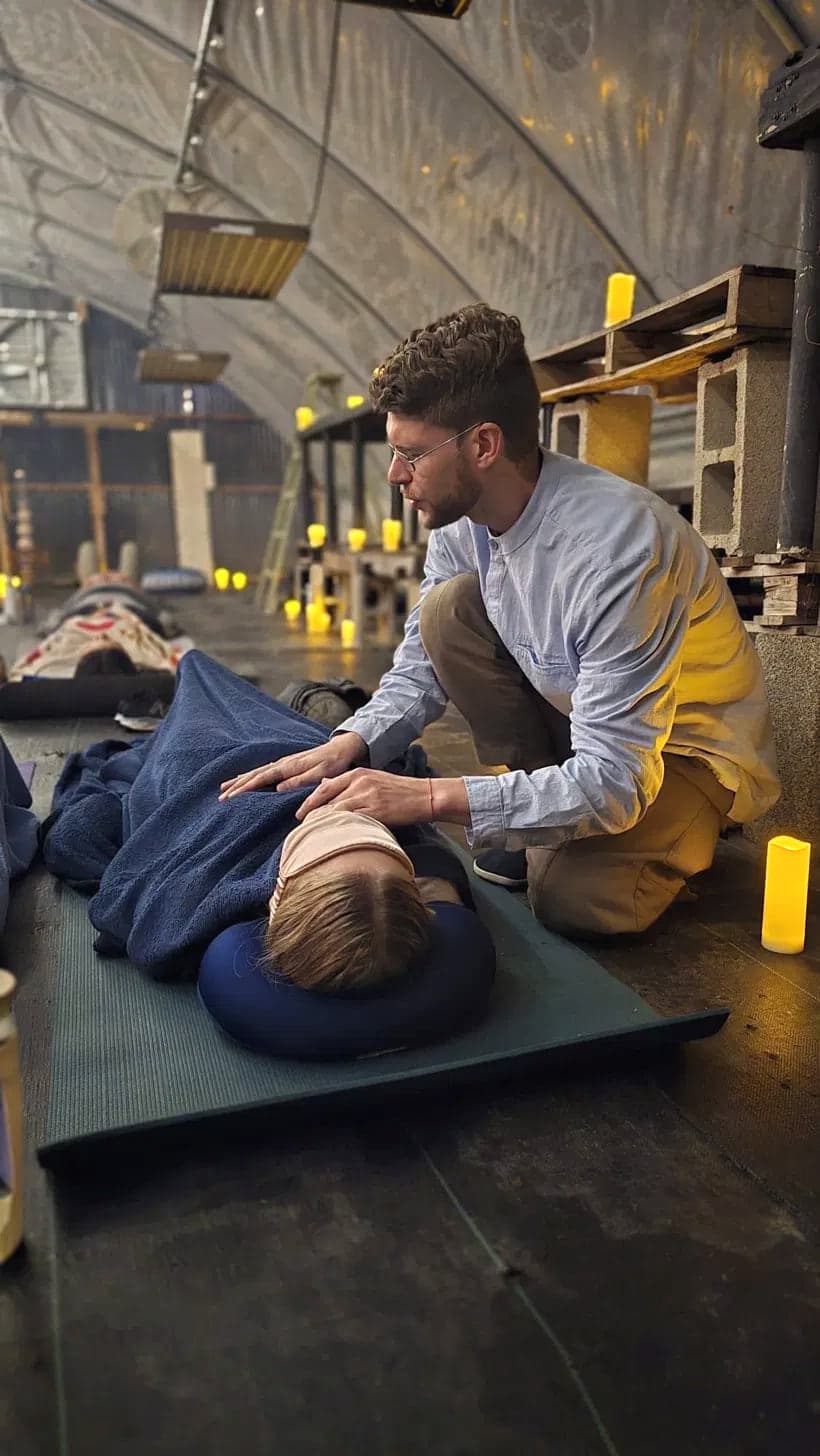 David Sauvé kneeling with one hand resting gently on the back of a person resting under a blanket, warm candlelight around them