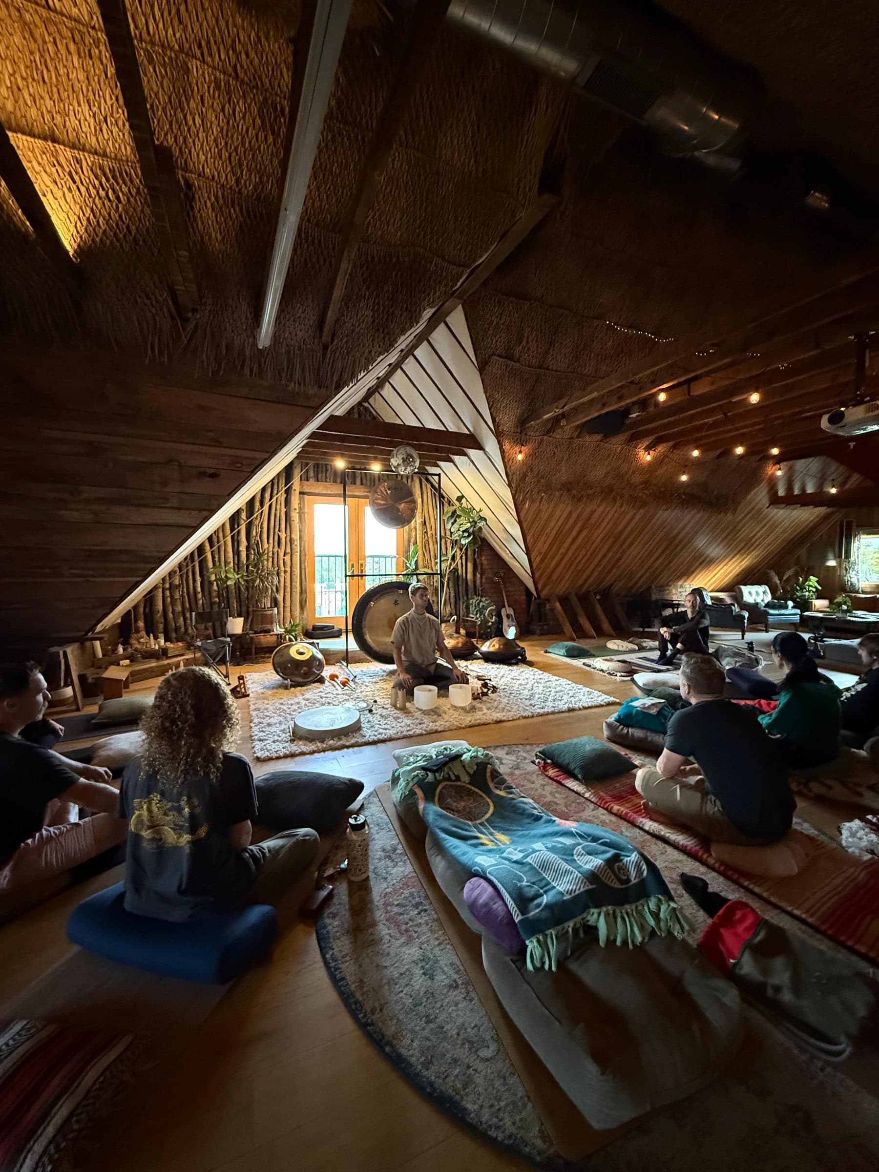 A group gathered in the A-frame sound space — David at the center with gongs, handpans, and singing bowls, people seated in a circle on floor cushions